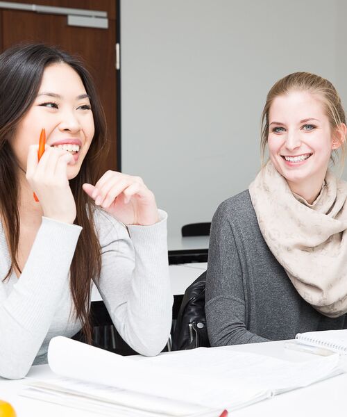 Zwei junge Frauen sitzen lachend mit Schreibunterlagen in einem Seminarraum.