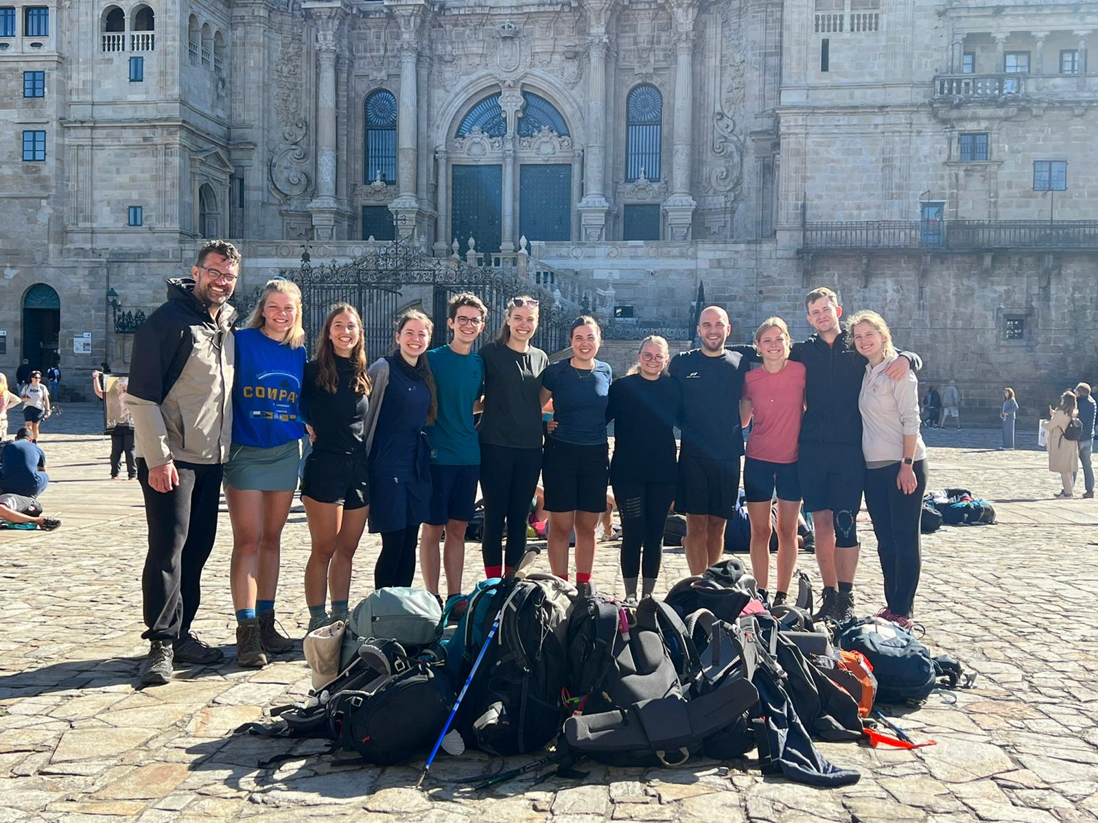 Gruppe von Menschen steht Arm in Arm auf einem Platz vor einer großen Kathedrale, im Vordergrund liegen zahlreiche Rucksäcke und Wanderstöcke.Gruppe von Menschen steht Arm in Arm auf einem Platz vor einer großen Kathedrale, im Vordergrund liegen zahlreich