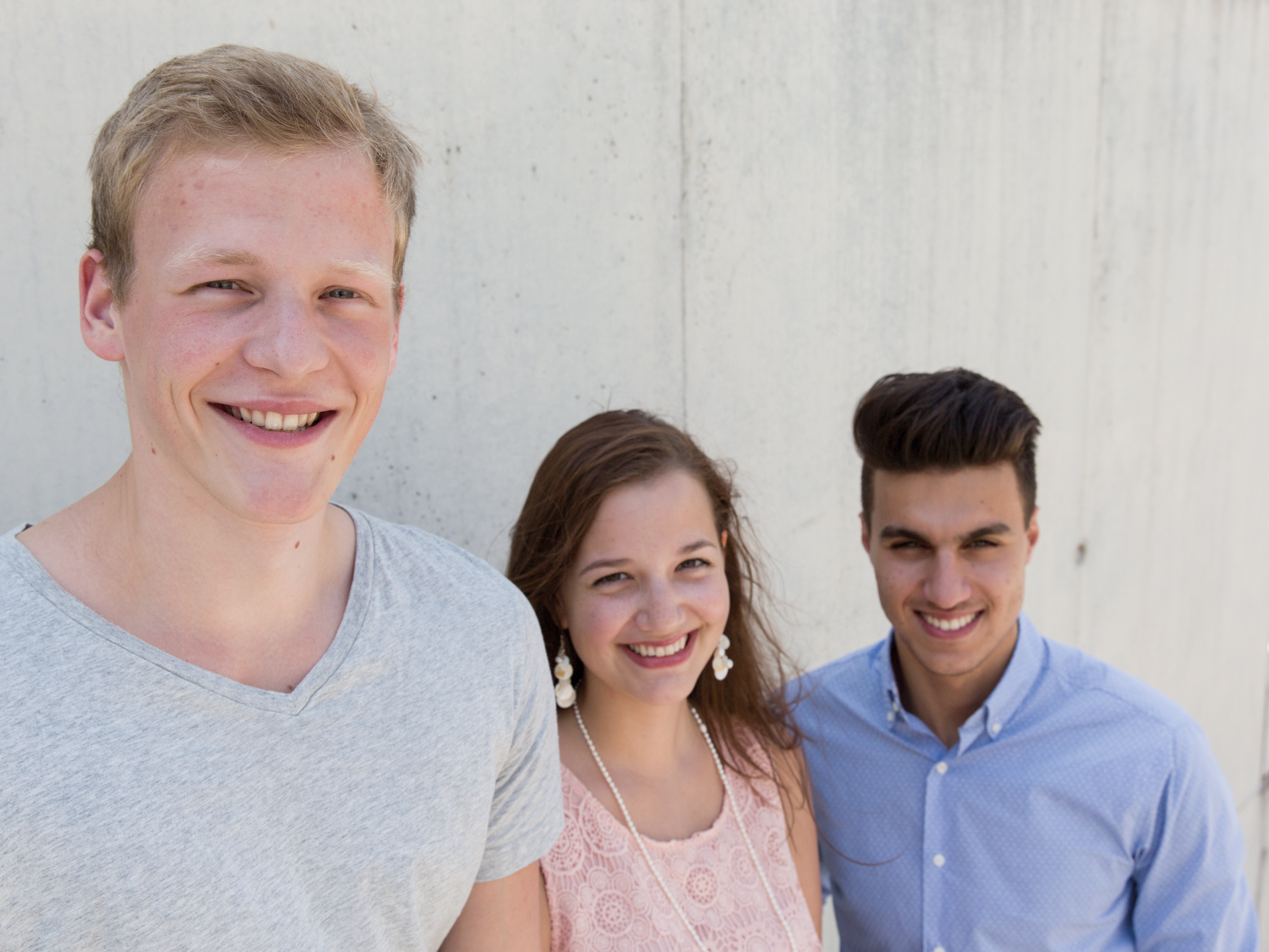 Drei junge Menschen stehen lächelnd nebeneinander vor einer hellen Betonwand.
