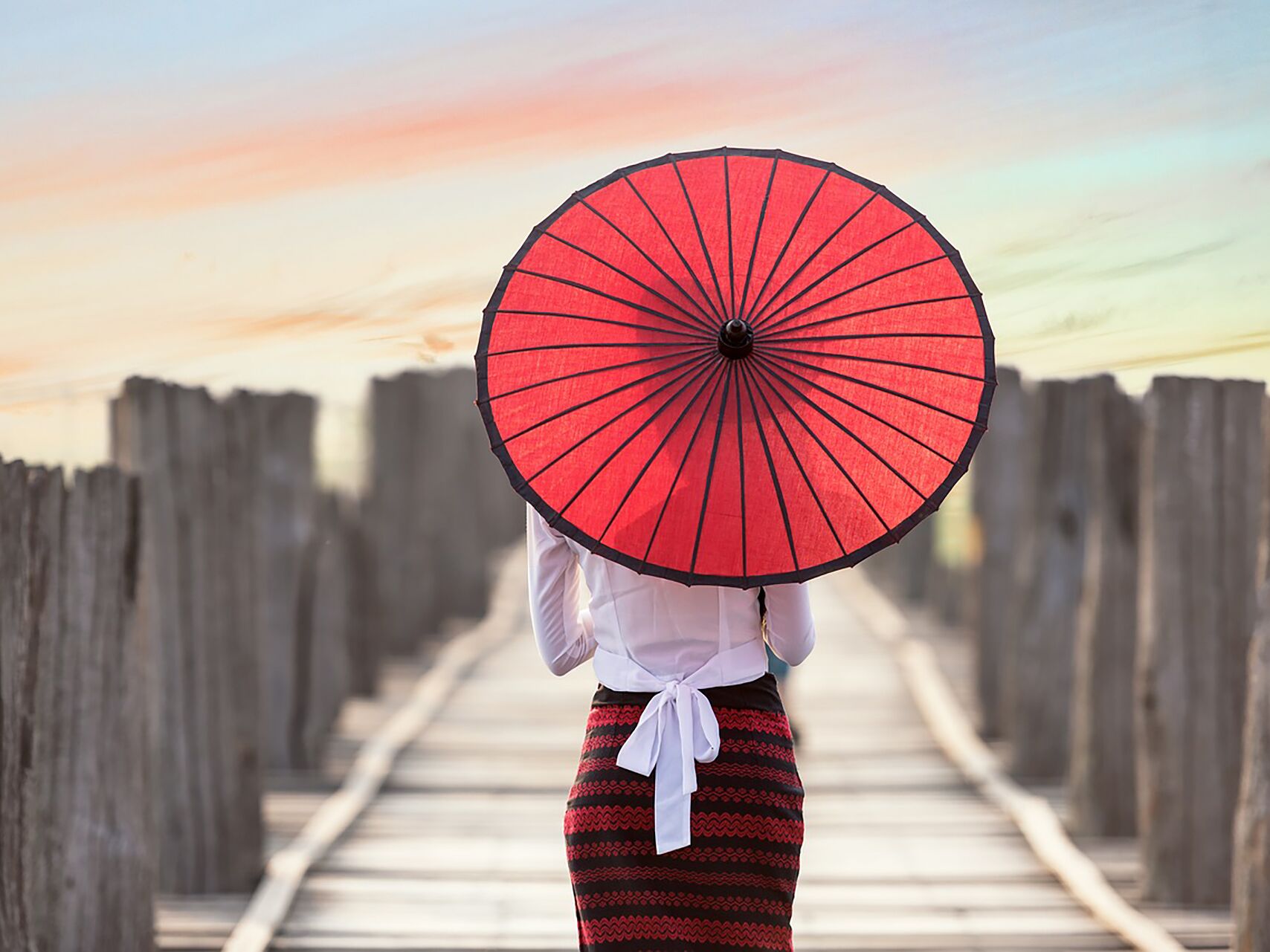 Person mit rotem Sonnenschirm steht auf einem Holzsteg bei Sonnenuntergang.