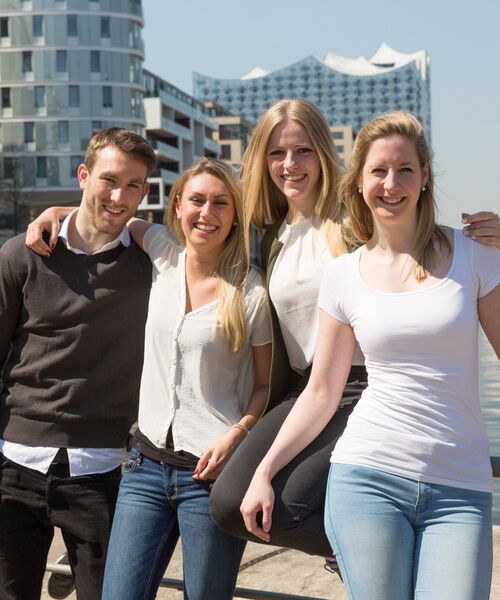 Vier junge Erwachsene stehen lachend zusammen im Freien vor moderner Architektur am Wasser.