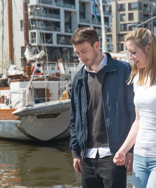Junger Mann und junge Frau gehen Hand in Hand am Wasser, im Hintergrund liegen Segelboote und moderne Gebäude.