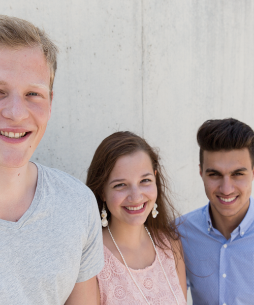 Drei junge Menschen stehen lächelnd nebeneinander vor einer hellen Betonwand.