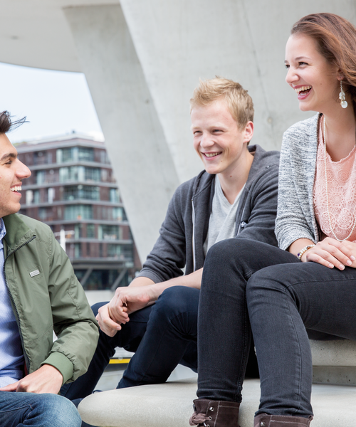 Drei junge Menschen sitzen draußen zusammen und lachen, im Hintergrund moderne Architektur.