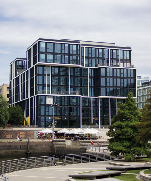 Modernes Bürogebäude mit Glasfassade am Wasser, davor eine Terrasse mit Sonnenschirmen.