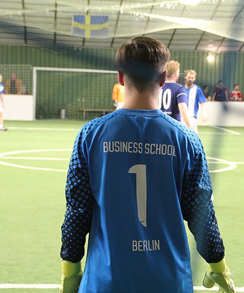 Hallenfußballspiel, ein Torwart im blauen Trikot mit der Aufschrift „Business School Berlin“ beobachtet das Spielgeschehen vor sich.