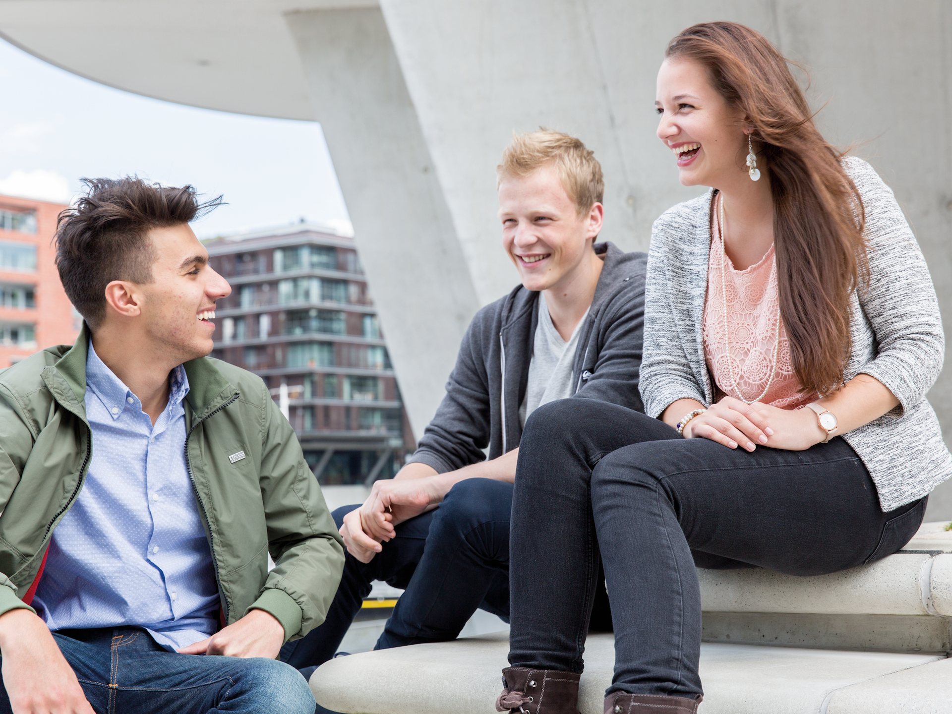 Drei junge Menschen sitzen draußen zusammen und lachen, im Hintergrund moderne Architektur.