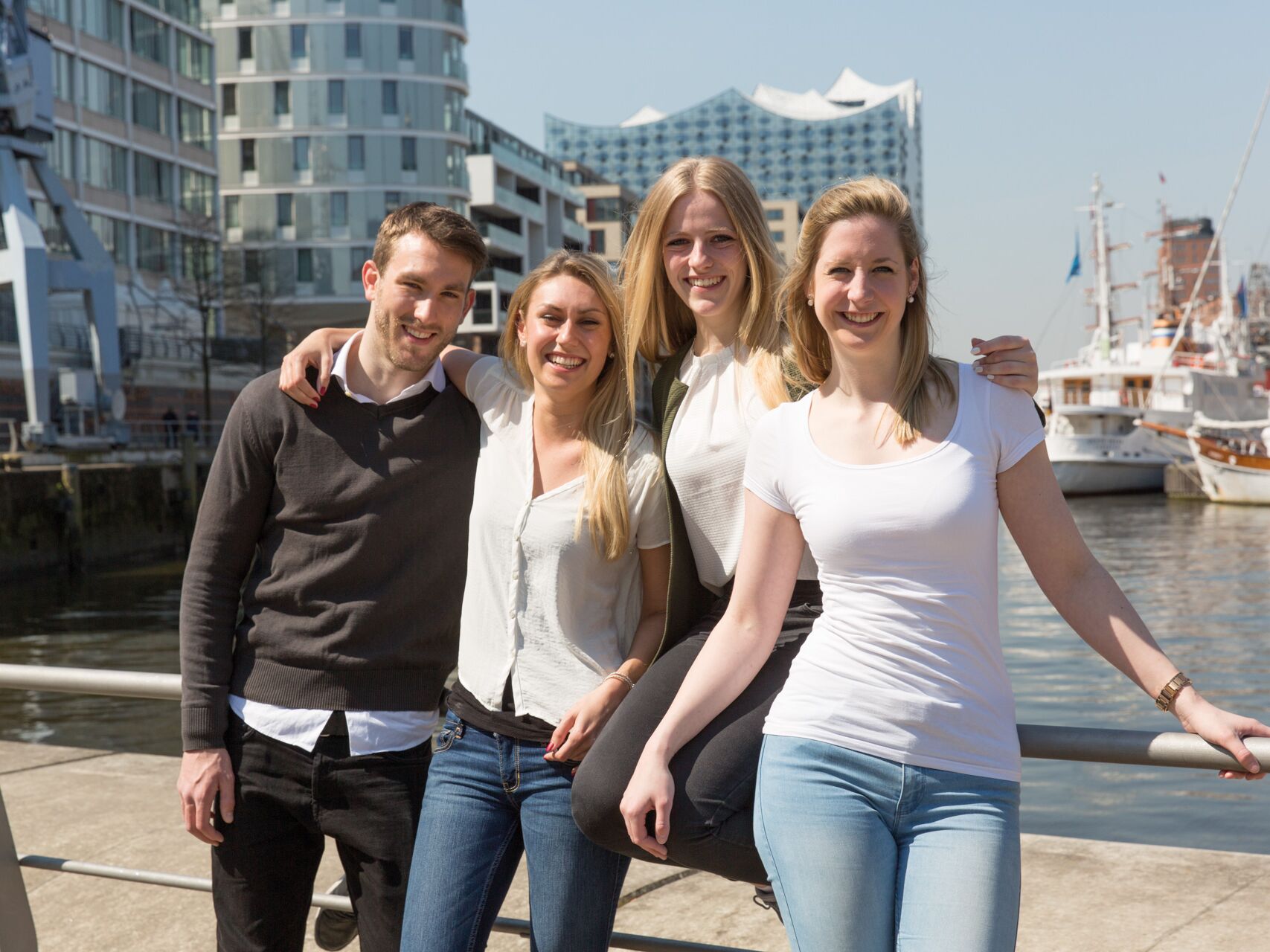 Vier junge Erwachsene stehen lachend zusammen im Freien vor moderner Architektur am Wasser.