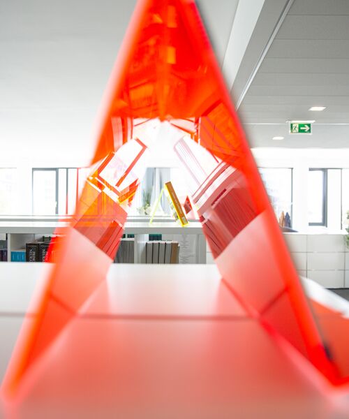 Rotes Acrylelement in Form eines Dreiecks, durch das man in einen modernen Innenraum mit Büchern blickt.