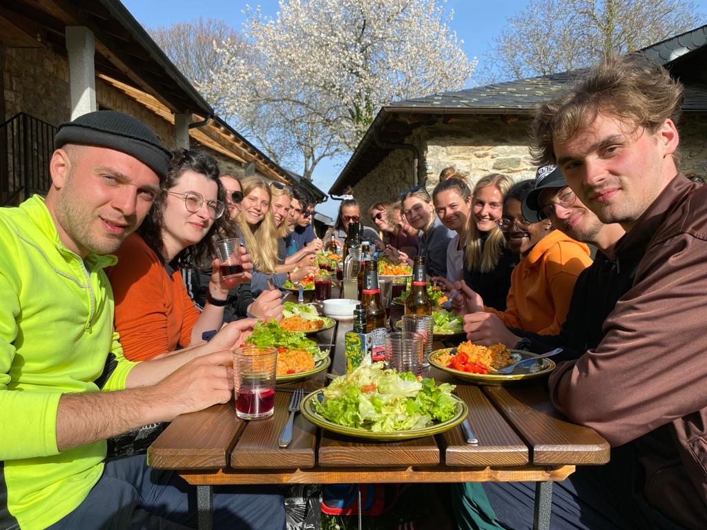 Eine große Gruppe junger Menschen sitzt draußen an einem langen Holztisch und isst gemeinsam, mit bunten Tellern voller Salat und Reisgerichten; im Hintergrund blühende Bäume und alte Steinhäuser.