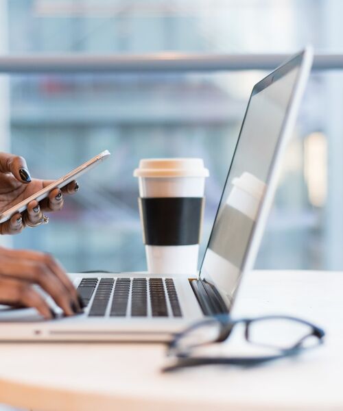 Person arbeitet am Laptop und hält ein Smartphone, daneben ein Kaffeebecher.
