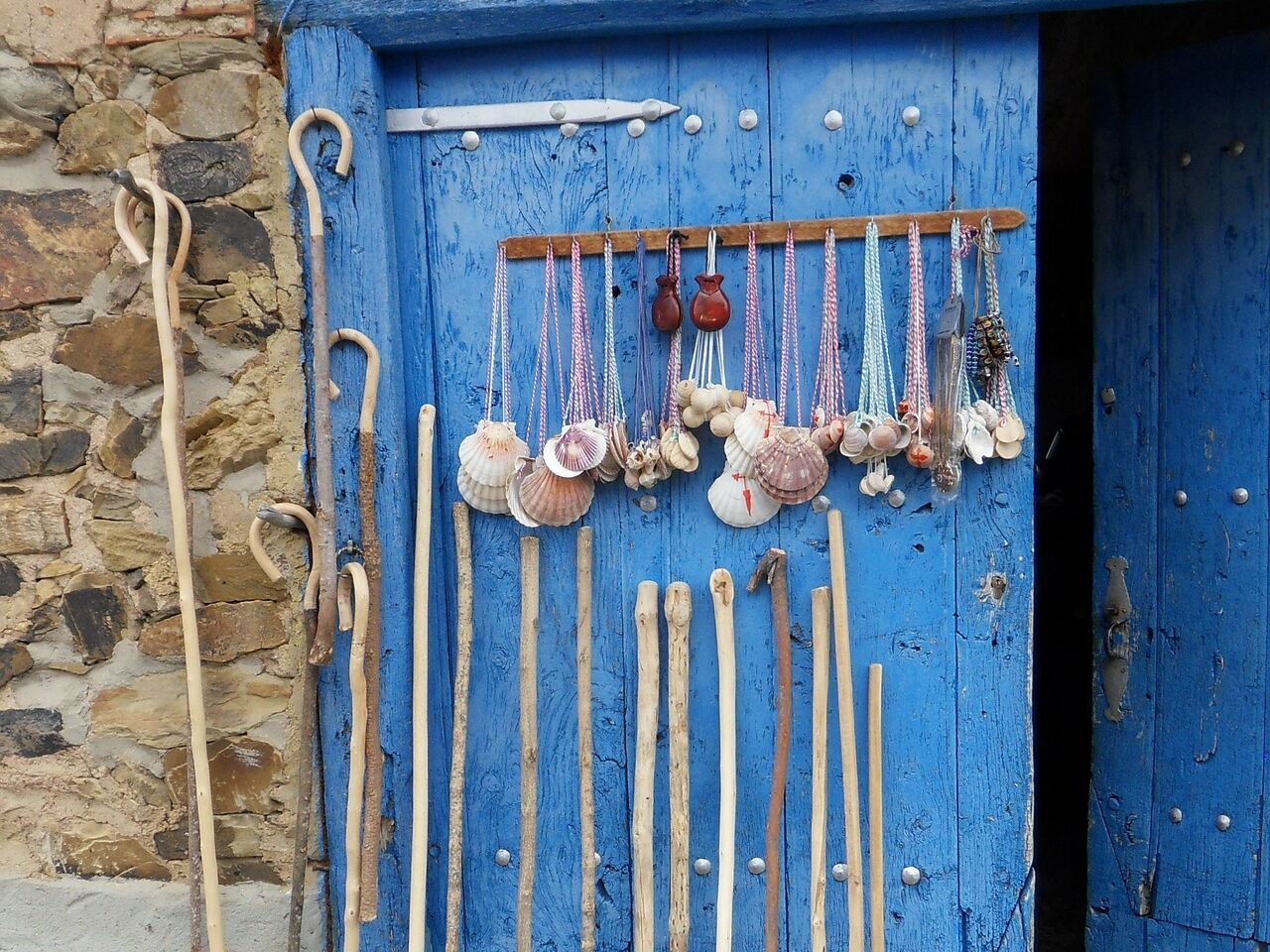 Blaue Holztür, an der Pilgerstäbe und Jakobsmuscheln mit bunten Bändern hängen, typisches Symbol und Ausrüstung des Jakobswegs.