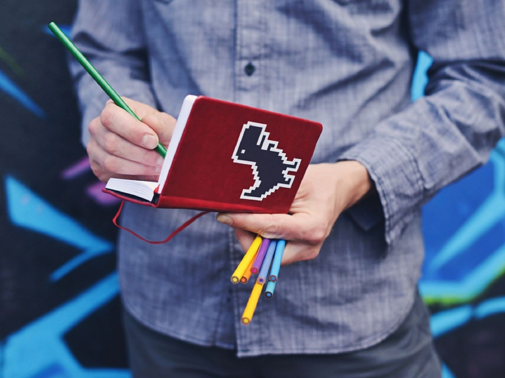 Person hält ein rotes Notizbuch mit Pixel-Art-Aufkleber eines Dinosauriers und schreibt mit einem grünen Buntstift, während sie mehrere bunte Stifte in der Hand trägt.