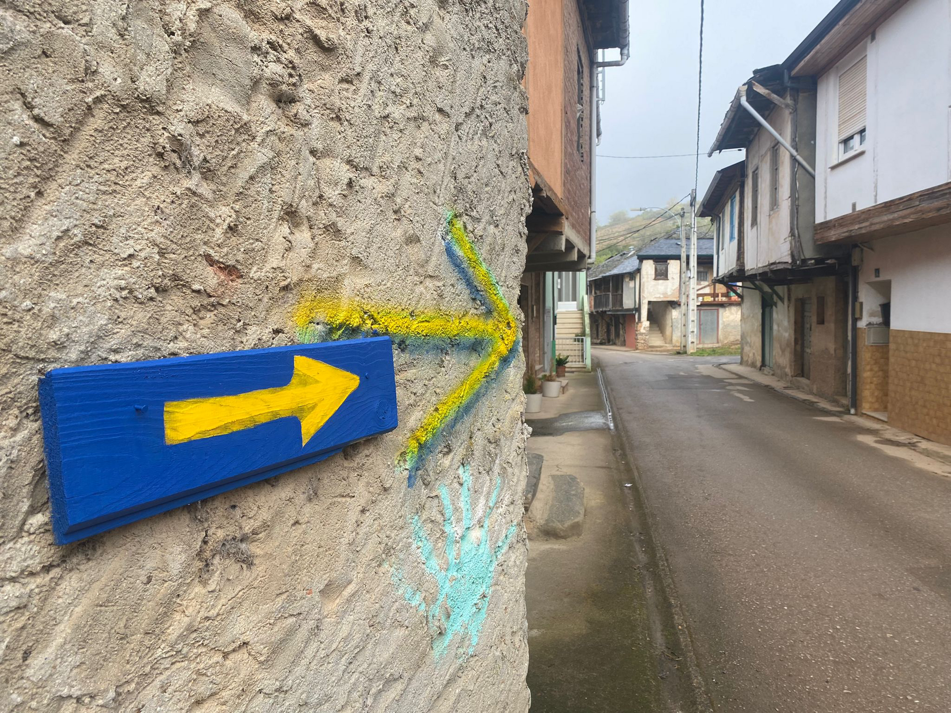 Blaues Holzschild mit gelbem Pfeil zeigt nach rechts, daneben ein gelber aufgesprühter Pfeil auf der Wand; im Hintergrund eine schmale Gasse mit alten Häusern.