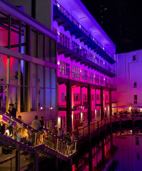 Modernes Gebäude am Wasser, nachts in pink-violettes Licht getaucht, mit Menschen auf der Terrasse.