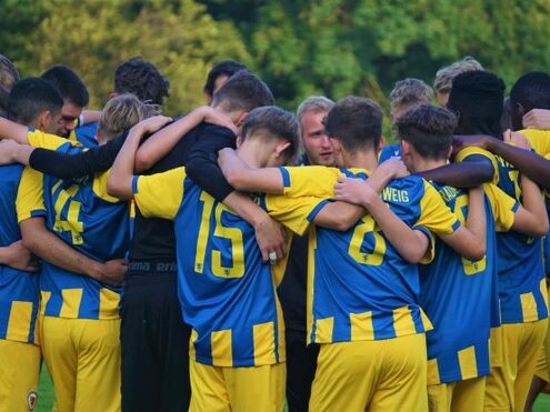 Eine Fußballmannschaft in blauen und gelben Trikots steht eng im Kreis zusammen, die Arme auf den Schultern der Teamkollegen. In der Mitte spricht eine Person, vermutlich der Trainer, zu den Spielern. Im Hintergrund ist grün bewaldete Natur zu sehen.