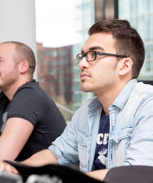 Junger Mann mit Brille und Jeanshemd sitzt aufmerksam in einem Seminarraum.