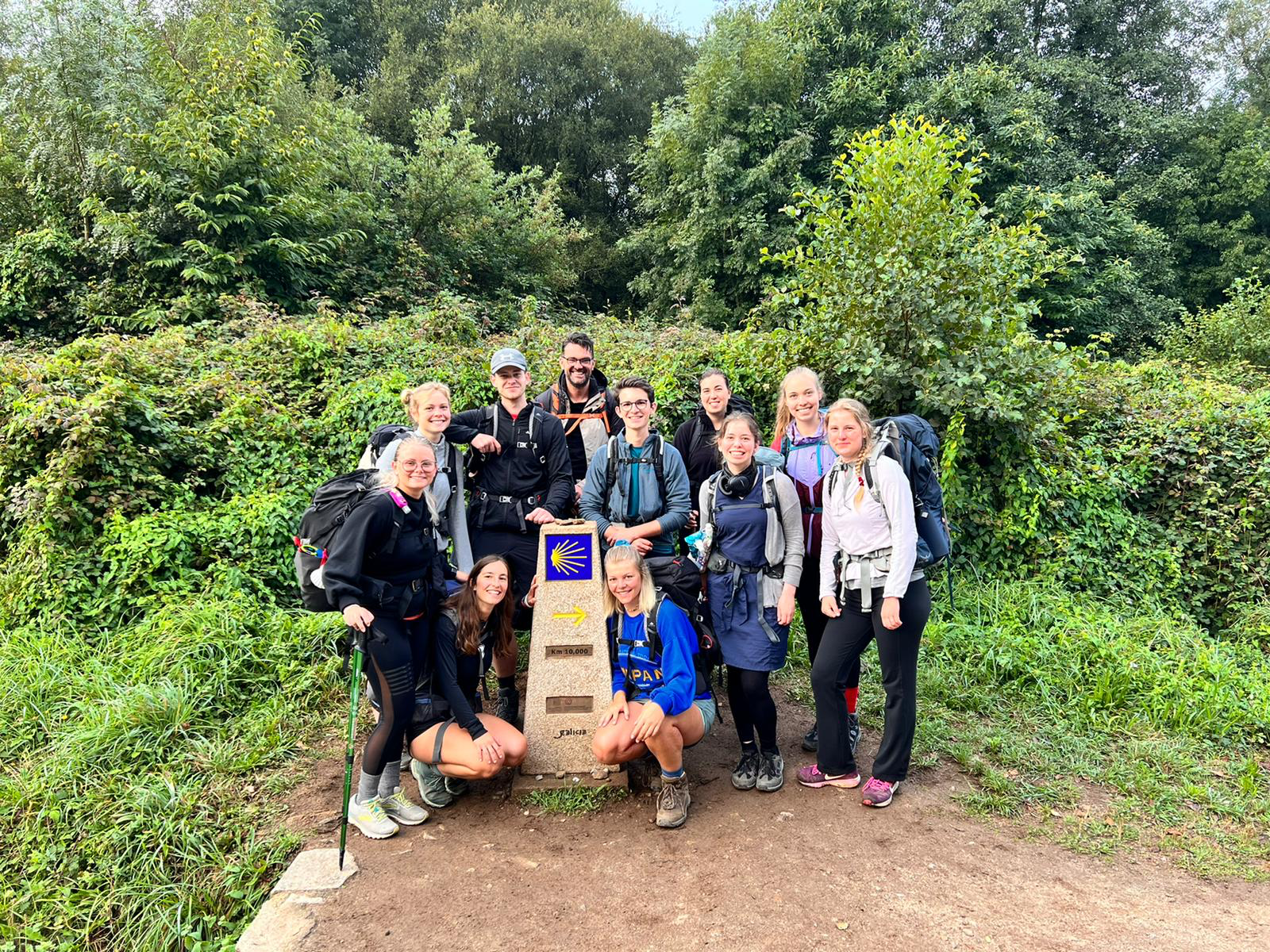 Gruppe von Wandernden mit Rucksäcken posiert fröhlich um einen Pilgerwegstein des Jakobswegs mit der gelben Muschel und einem Pfeil, im Hintergrund dichte grüne Vegetation.