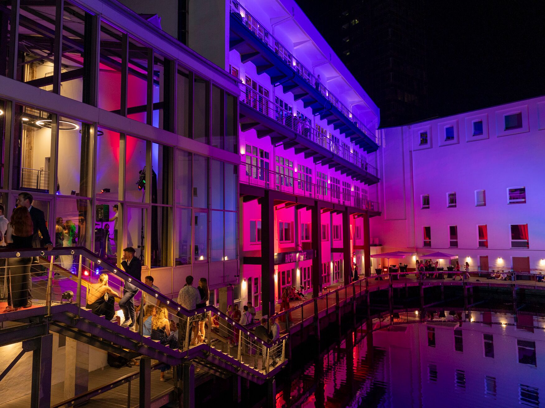 Modernes Gebäude am Wasser, nachts in pink-violettes Licht getaucht, mit Menschen auf der Terrasse.