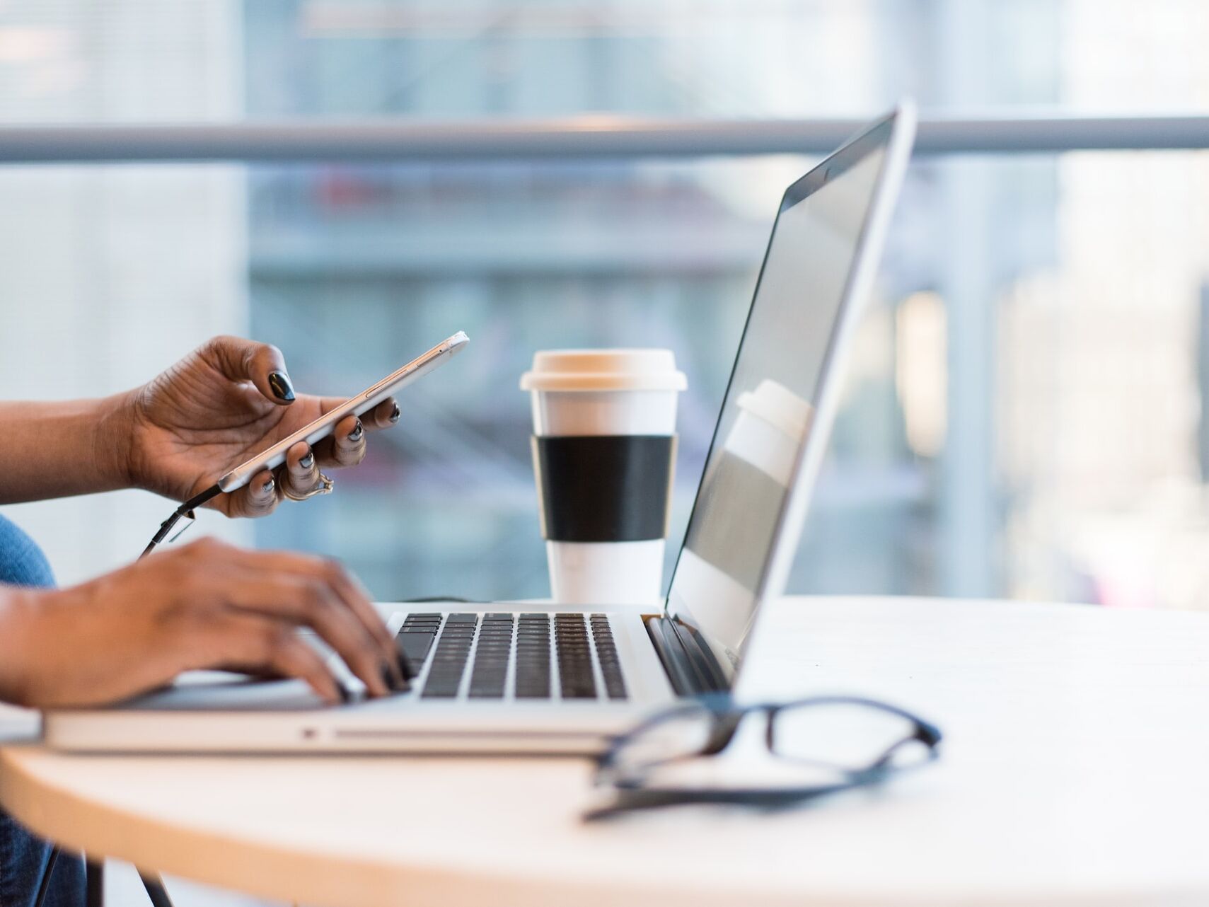 Person arbeitet am Laptop und hält ein Smartphone, daneben ein Kaffeebecher.