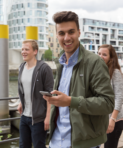 Drei junge Erwachsene spazieren lachend am Wasser entlang, einer hält ein Smartphone in der Hand.
