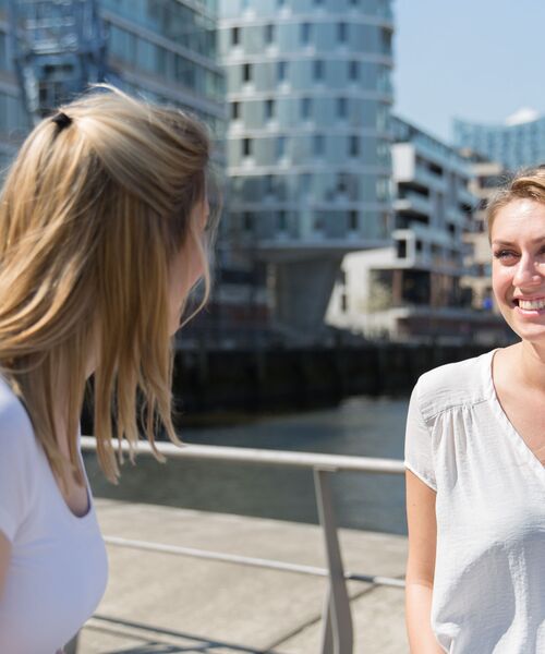 Zwei Frauen unterhalten sich lachend im Freien vor moderner Architektur am Wasser.