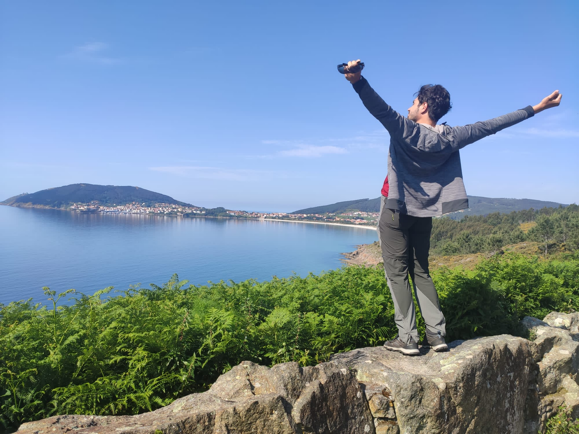 Person steht mit ausgebreiteten Armen auf einem Felsen und blickt über das Meer und eine Küstenstadt in der Ferne.