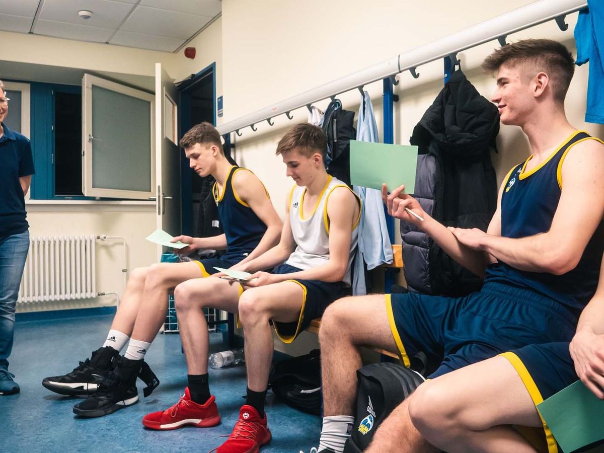 Junge Basketballspieler sitzen in einem Umkleideraum auf einer Bank, tragen Trainingskleidung und halten grüne Karten in der Hand, während sie lächeln und miteinander sprechen.
