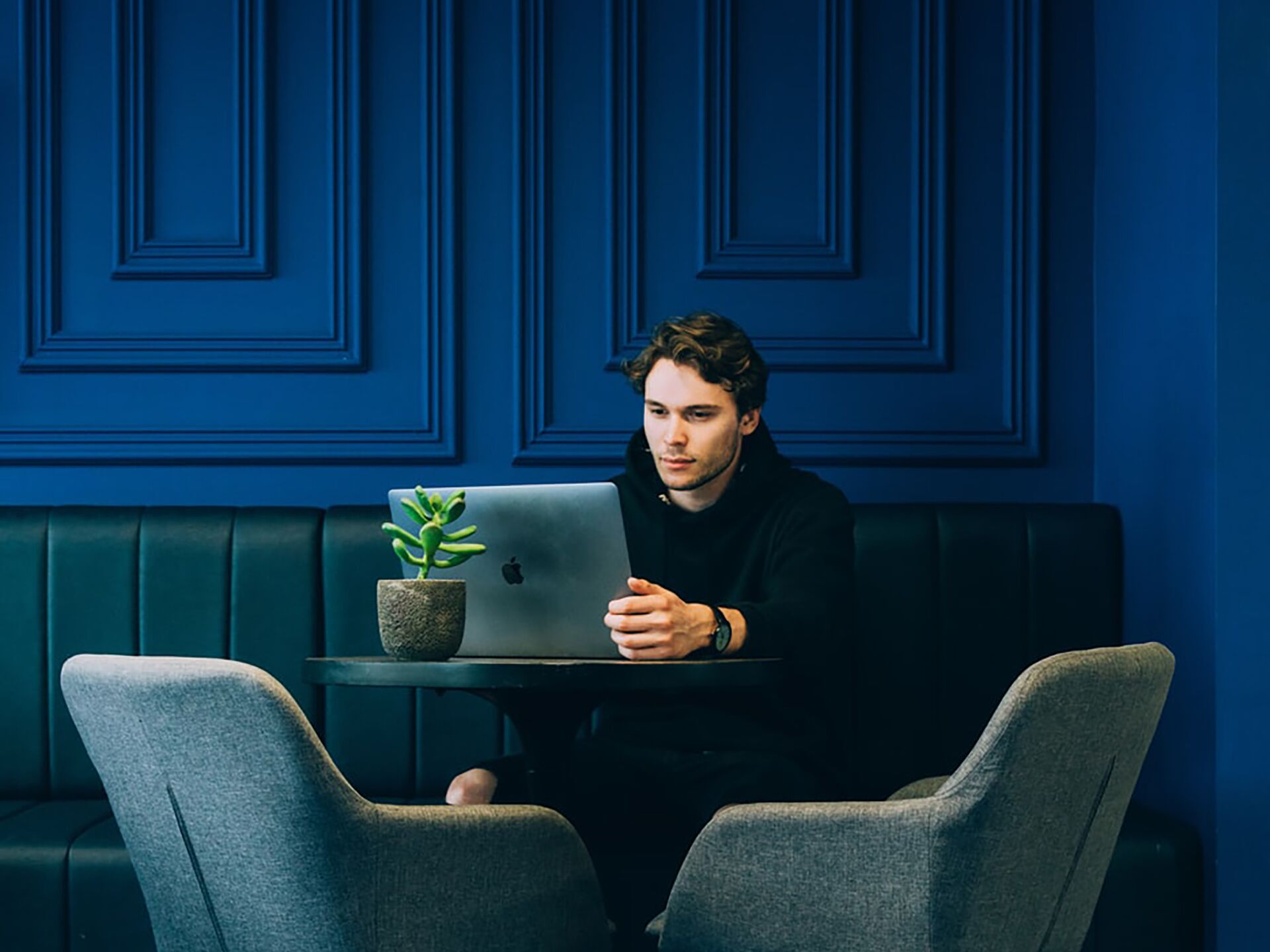 Junger Mann sitzt an einem kleinen Tisch mit Laptop in modern eingerichteter Lounge mit blauen Wänden.