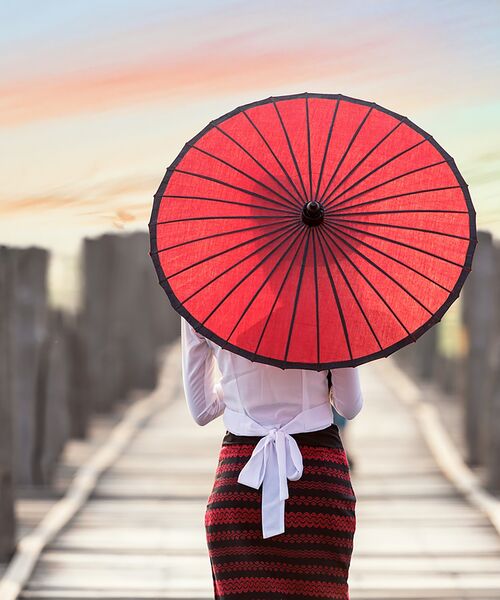 Person mit rotem Sonnenschirm steht auf einem Holzsteg bei Sonnenuntergang.