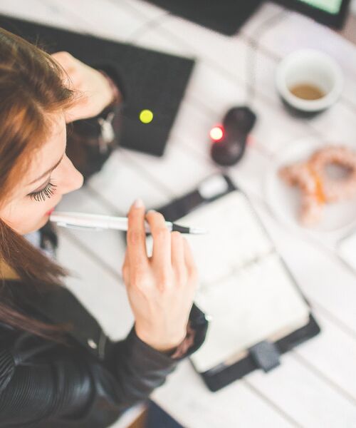 Frau sitzt am Schreibtisch, hält einen Stift und blickt nachdenklich in einen offenen Planer.
