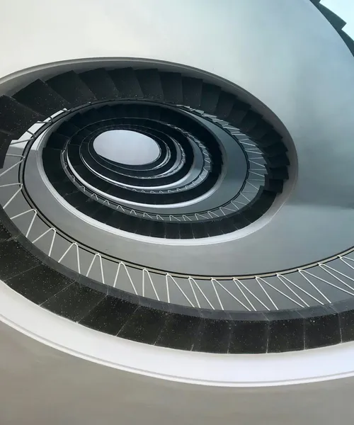 Blick nach oben in ein spiralförmiges Treppenhaus mit schwarzen Stufen und weißen Geländern.