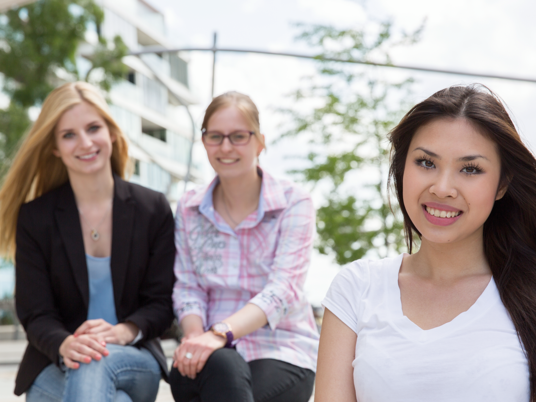 Drei junge Frauen sitzen draußen zusammen, eine im Vordergrund lächelt in die Kamera.