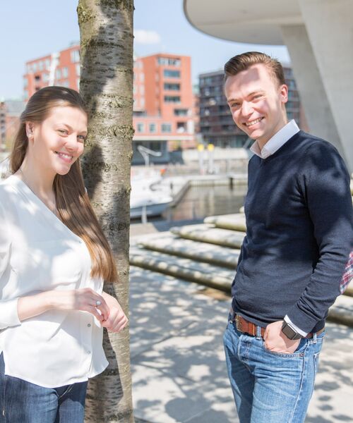Eine Frau und ein Mann stehen lachend im Freien an einem Baum, im Hintergrund moderne Architektur am Wasser.
