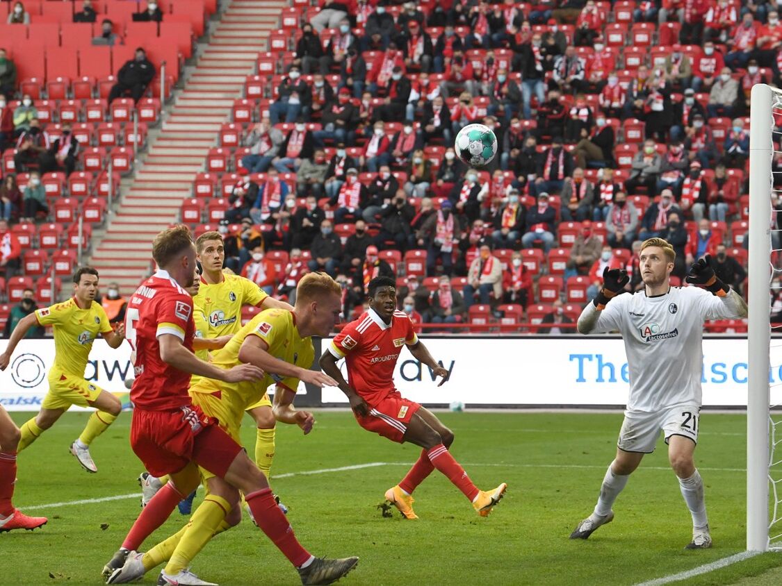 Fußballspiel in einem Stadion – Spieler in roten und gelben Trikots kämpfen um den Ball vor dem Tor, während der Torwart in weißer Ausrüstung die Situation beobachtet. Im Hintergrund sitzen Fans in rot-weißen Schals auf den Tribünen.