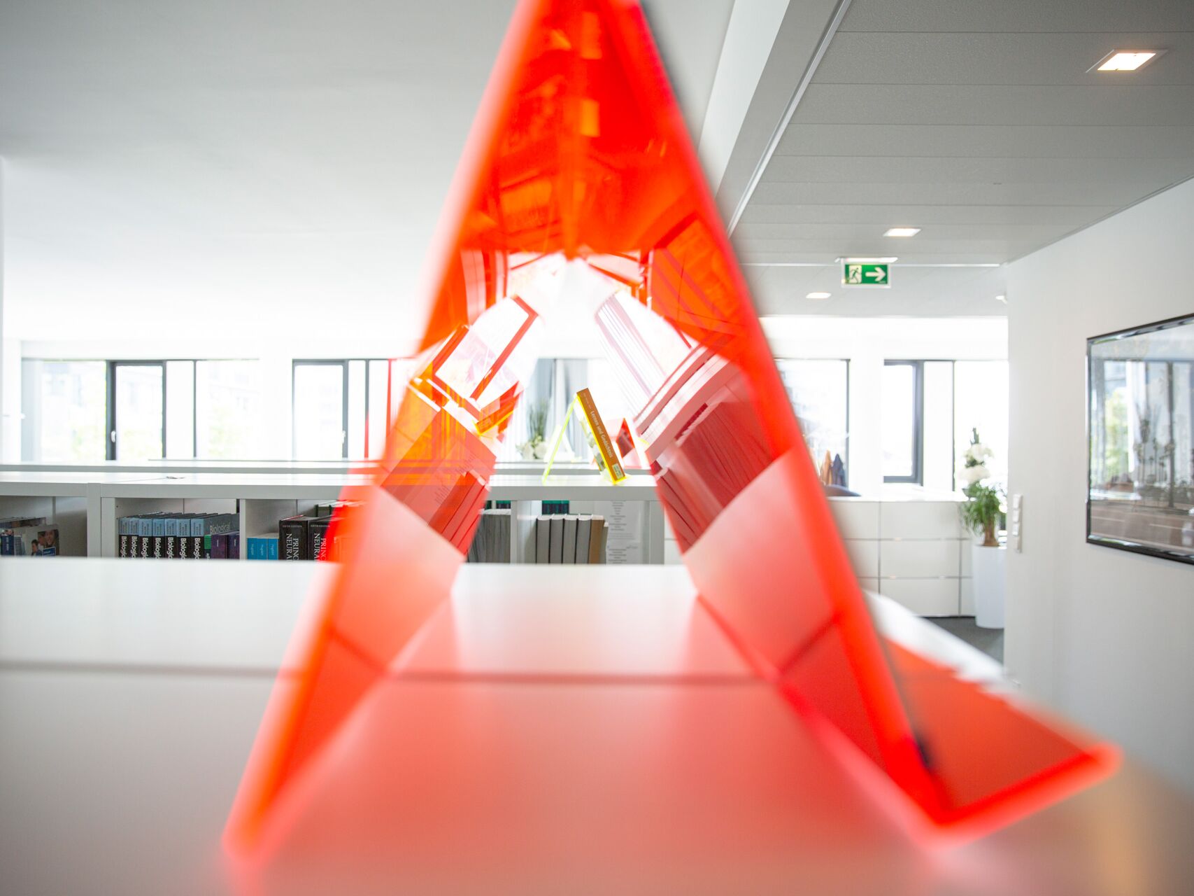 Rotes Acrylelement in Form eines Dreiecks, durch das man in einen modernen Innenraum mit Büchern blickt.