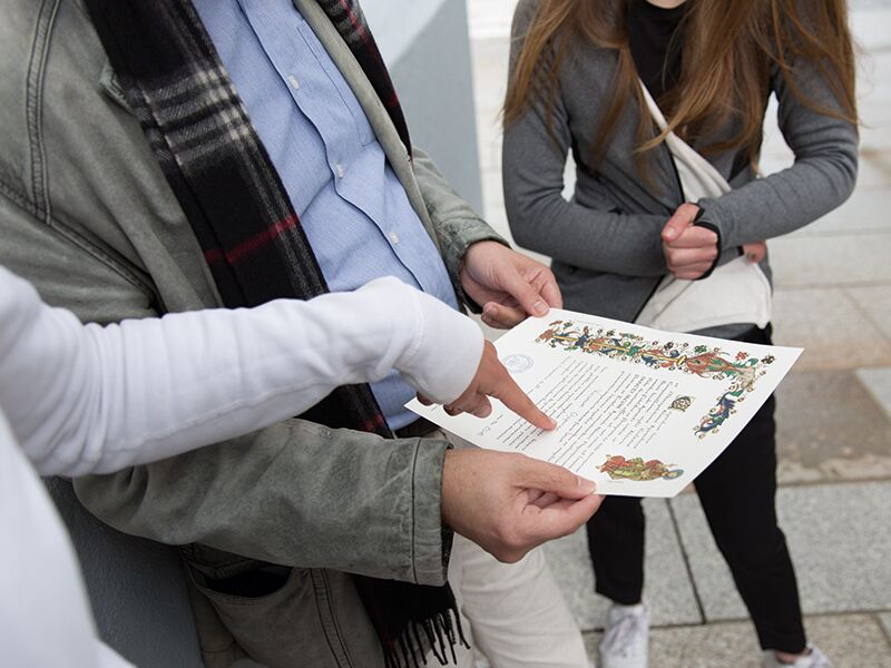 Eine Person hält eine verzierte Urkunde mit farbigen Illustrationen in den Händen, während eine andere darauf zeigt und eine dritte danebensteht.