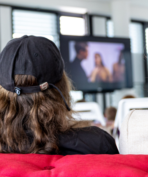 Person mit schwarzer Kappe sitzt auf einem roten Sofa und schaut auf einen Bildschirm im Hintergrund.