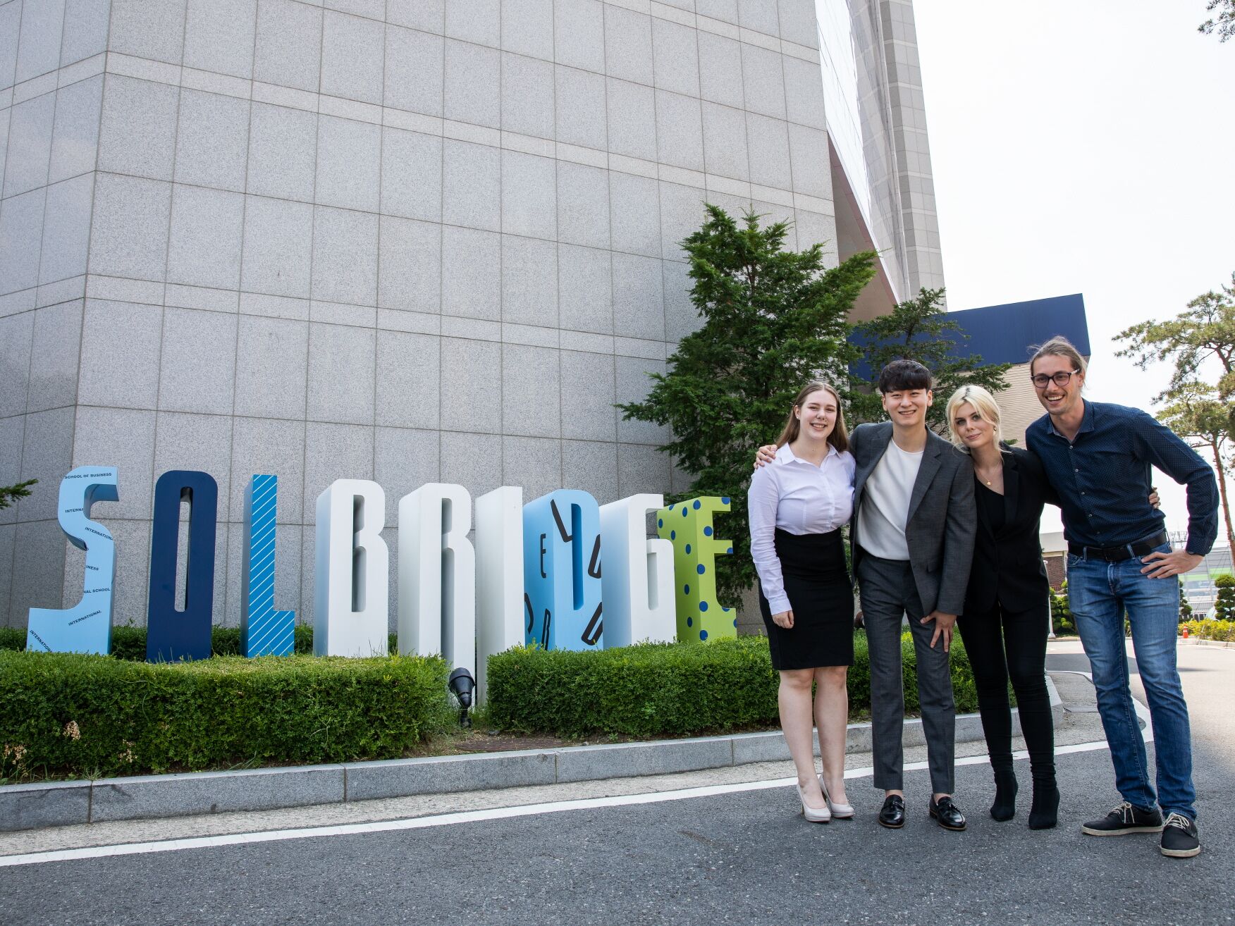 Vier Personen stehen lächelnd vor einem Gebäude neben großen bunten Buchstaben mit der Aufschrift SolBridge.