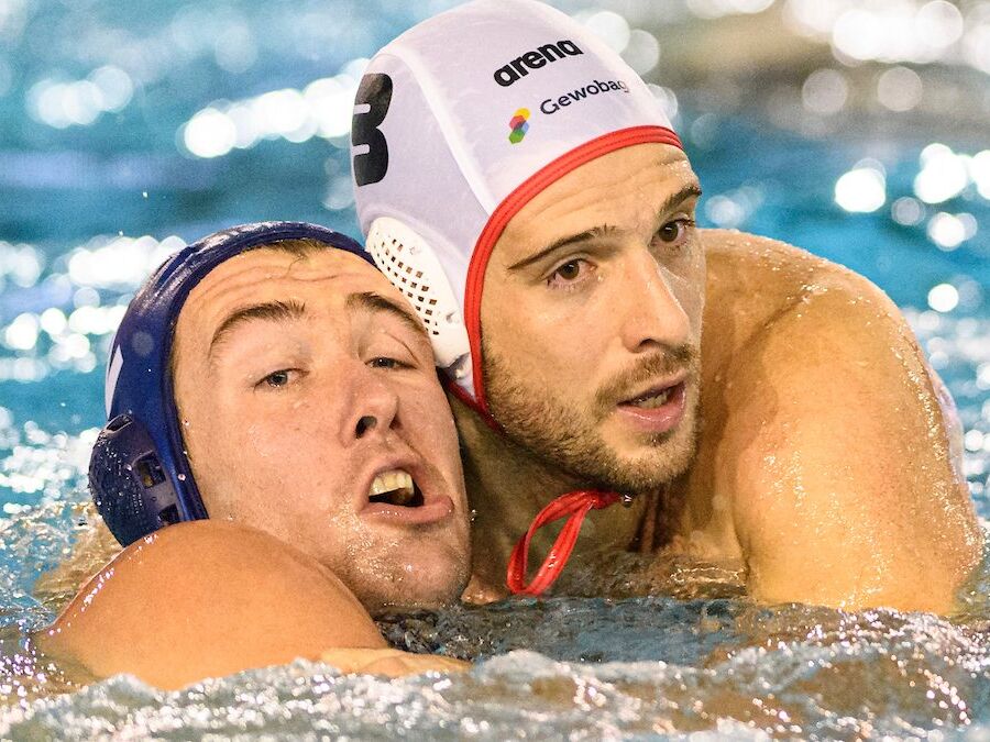 Zwei Wasserballspieler kämpfen im Wasser um Position. Der Spieler links trägt eine blaue Kappe, der rechte Spieler eine weiße Kappe mit rotem Rand und der Nummer 3. Beide liegen eng beieinander im Wasser, während sie konzentriert nach vorne blicken. Im Hi