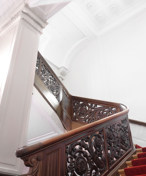 Ein prächtiges Treppengeländer aus dunkelbraunem Holz mit aufwendig geschnitzten Ornamenten führt eine Treppe mit rotem Teppich hinauf. Die helle weiße Wand und die verzierte Stuckdecke betonen den eleganten, historischen Charakter des Gebäudes.