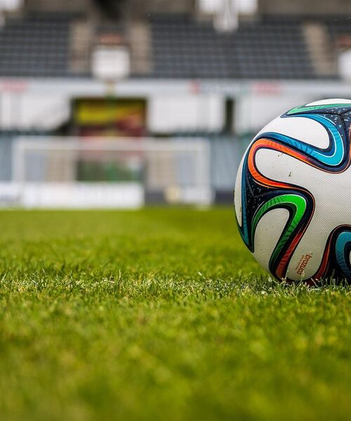 Ein Fußball liegt auf dem grünen Rasen eines Stadions, im Hintergrund sind verschwommen die Tribünen und ein Tor zu sehen.